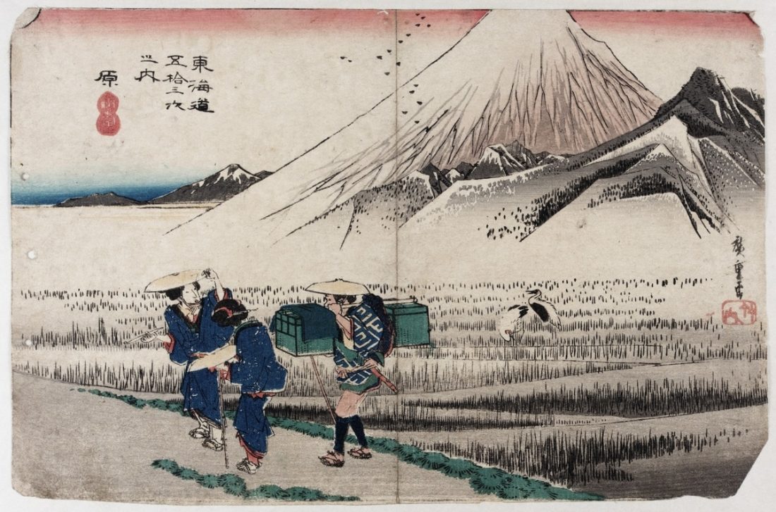 Japanese colour wood block print featuring a landscape scene of two female and one male figure walking through a snowy rice field below a crane bird and a snow topped Mount Fuji. This is one print from Hiroshige's Tokaido Road series.