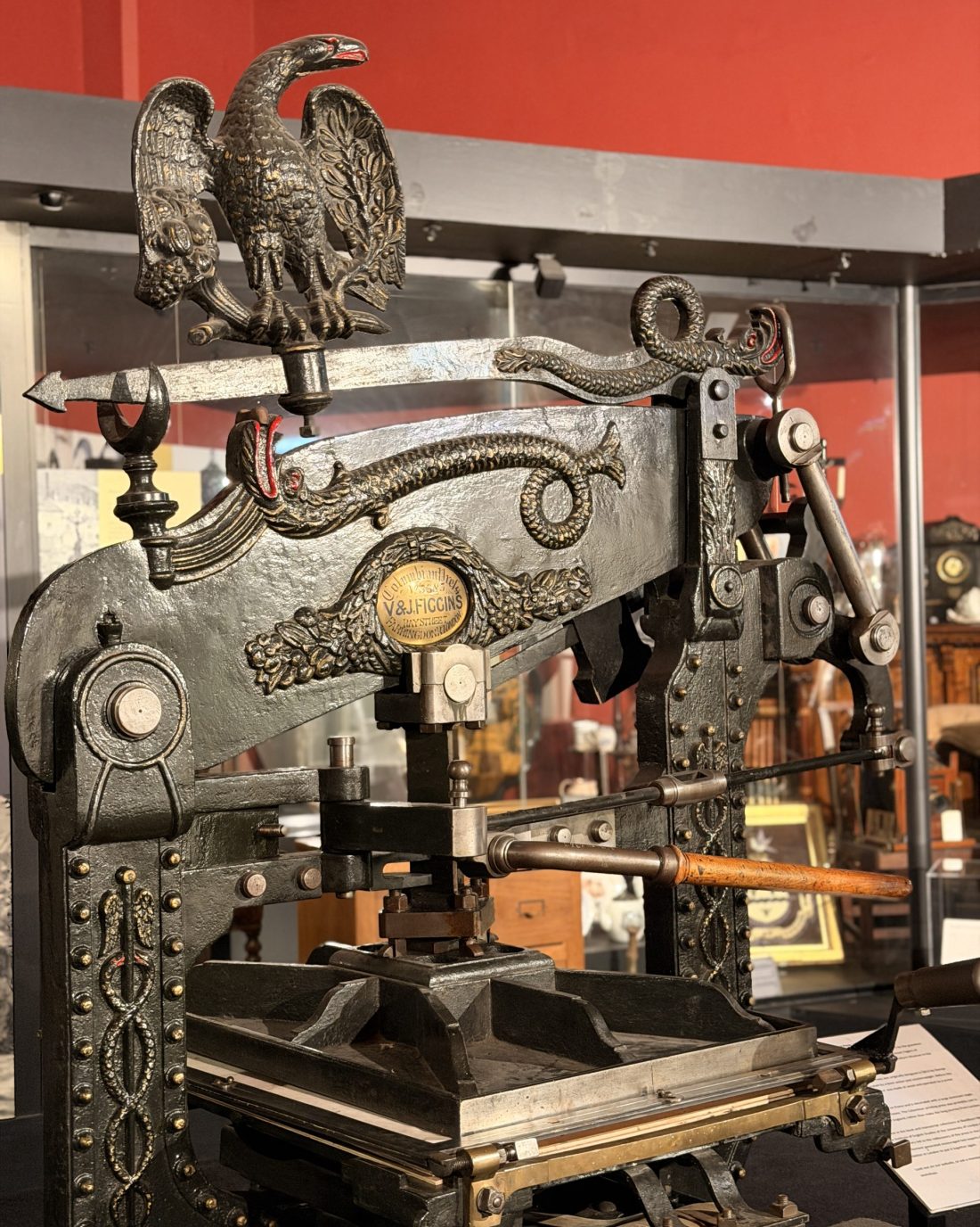 Large black cast iron printing press with a wooden handle. It is decorated with gold studs and designs. The top is adorned with an eagle sculpture and in the centre is a gold plaque with the makers name 'V&J Figgins'.