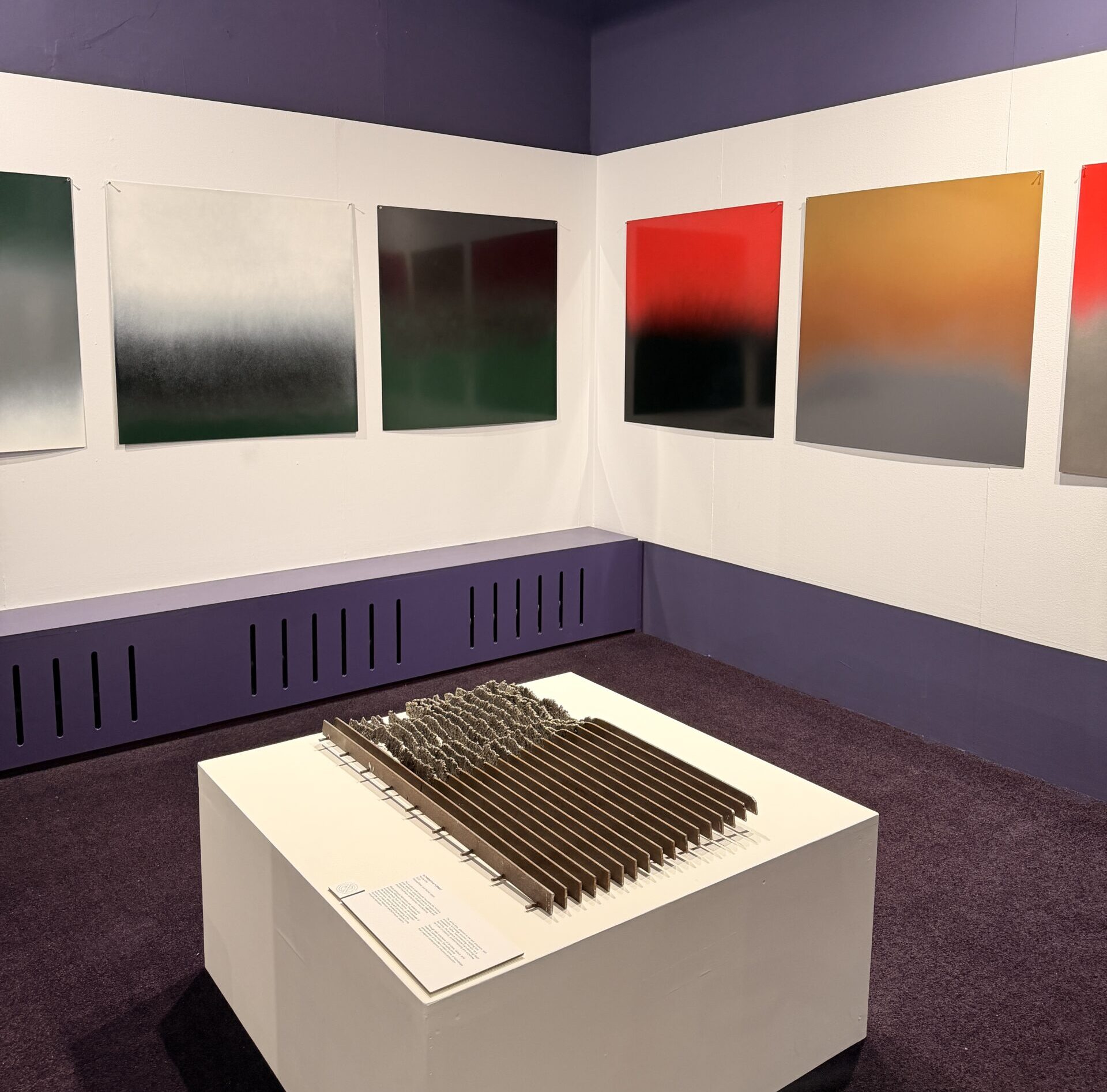 Hung one a white wall are 4 square metal sheet artworks decorated with colour gradients including black, white, green, red, orange and grey tones. In the centre is a metal railing displayed on a white plinth.