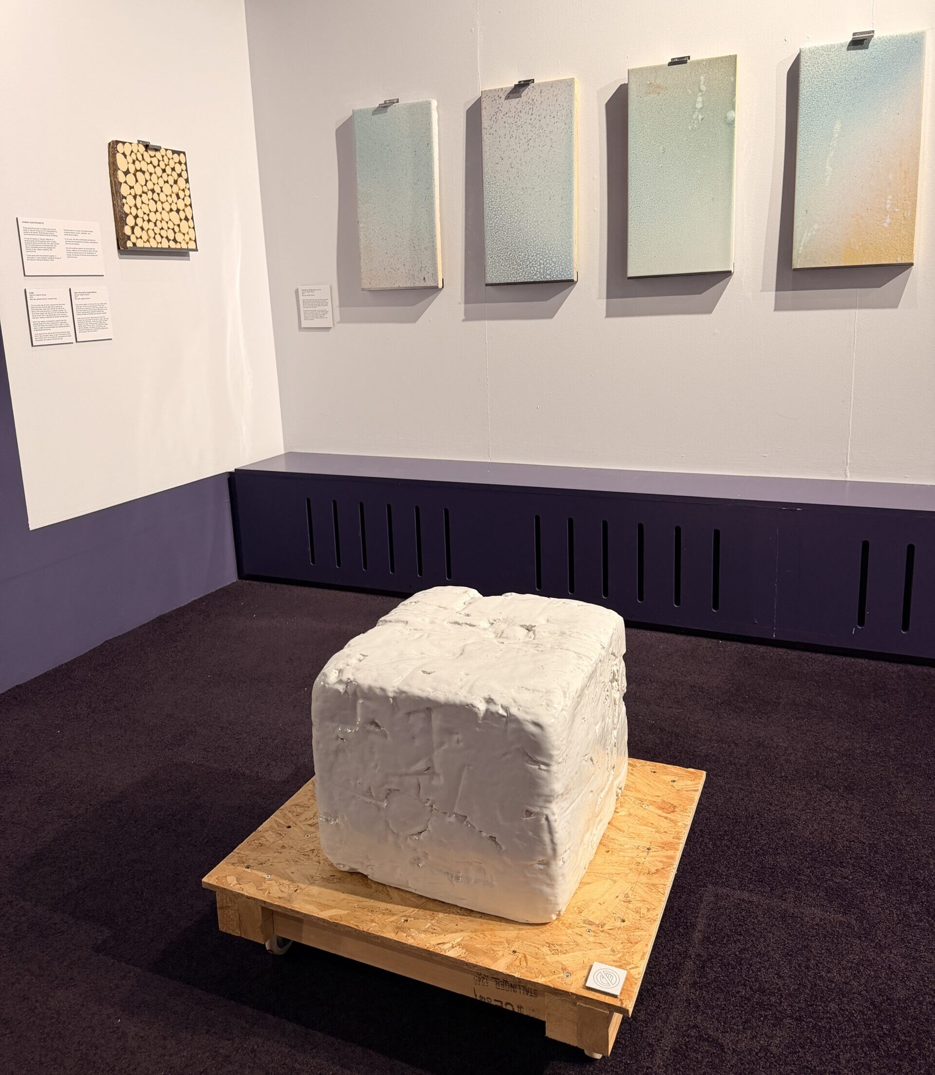 Hanging on a white wall is a square ceramic brown artwork next to 4 vertical rectangular pieces in pastel blue, green and cream tones. On the floor a white ceramic cube sits on a wooden plinth.