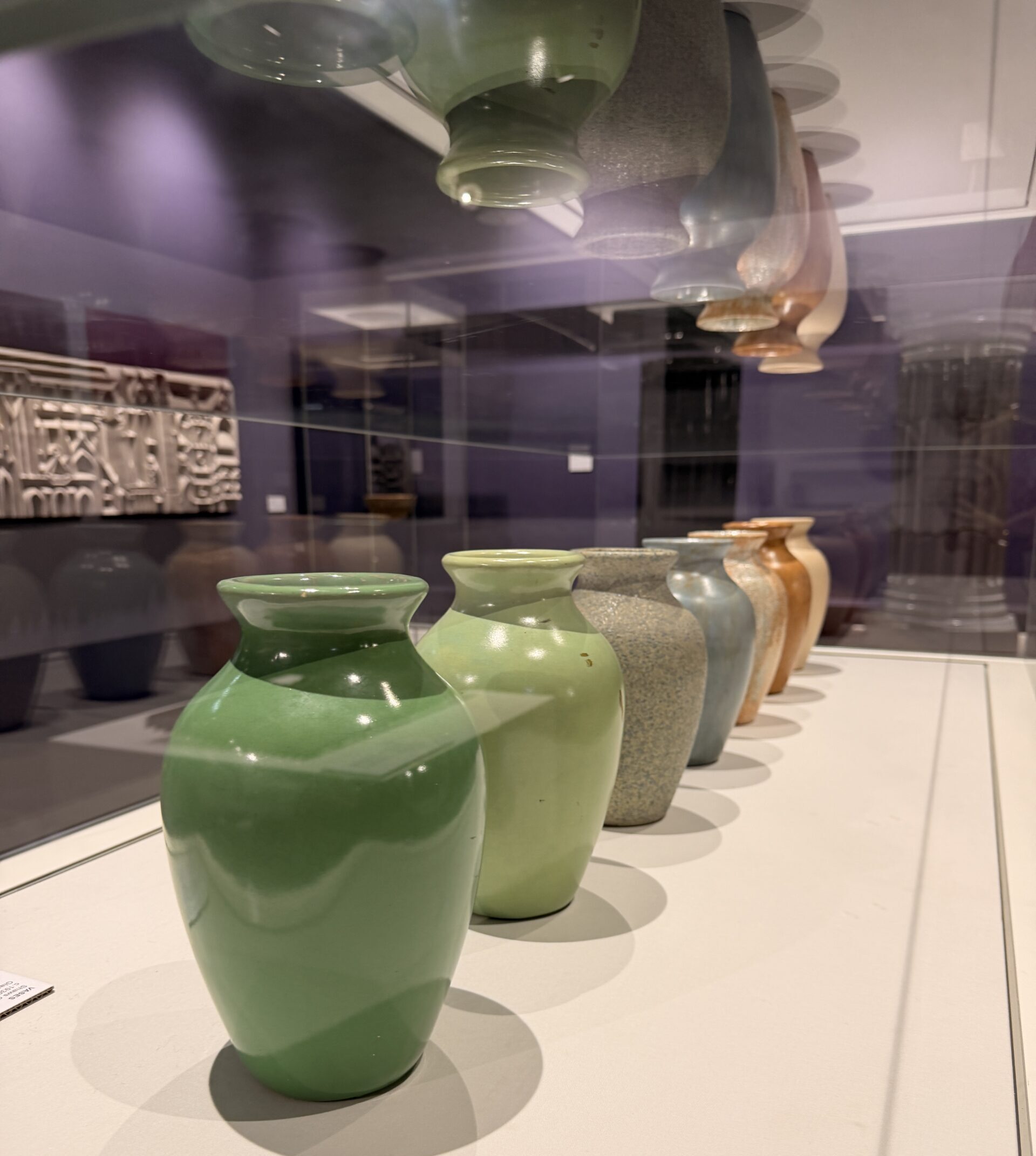 Inside a glass case 7 coloured vases of the same size and shape are displayed in a line. Above is a reflection of the vases caught in the glass. In the background is a wall mounted white artwork.